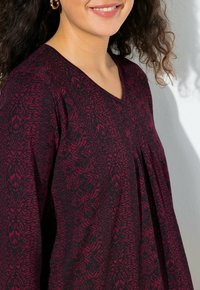 Haut à motifs rouge et noir avec un col en V, des manches longues et des détails plissés, fabriqué dans un tissu doux avec une finition texturée.