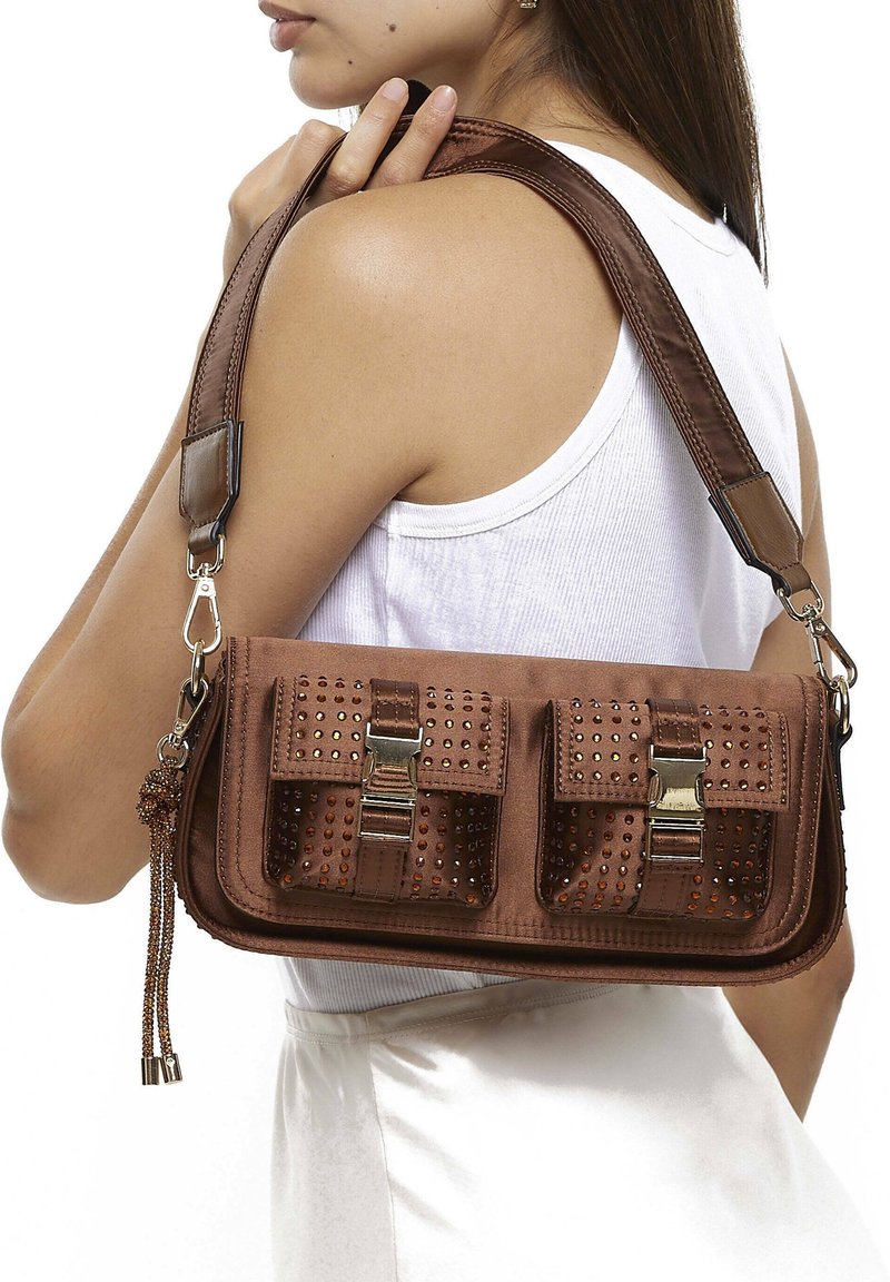 Femme portant un débardeur blanc porte un sac à bandoulière marron avec deux poches avant à boucle et des clous décoratifs.