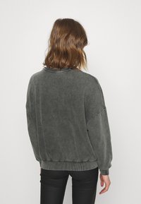 Person with shoulder-length brown hair wearing a washed dark gray long-sleeve sweater and black pants, facing away against a plain background.