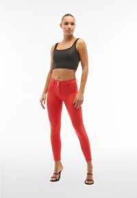 Top corto nero con zip, abbinato a leggings rossi a vita alta con tasche frontali e chiusura a doppio bottone, indossato con tacchi neri.