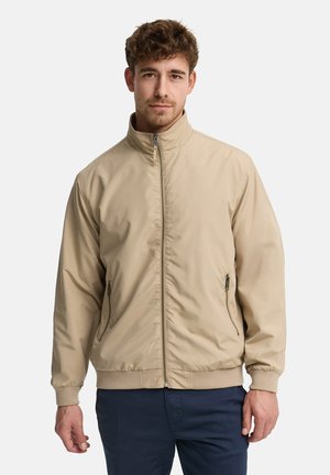 Mand iført beige lynlåstrøje med høj krave og marineblå bukser, stående mod en ensartet lys baggrund.