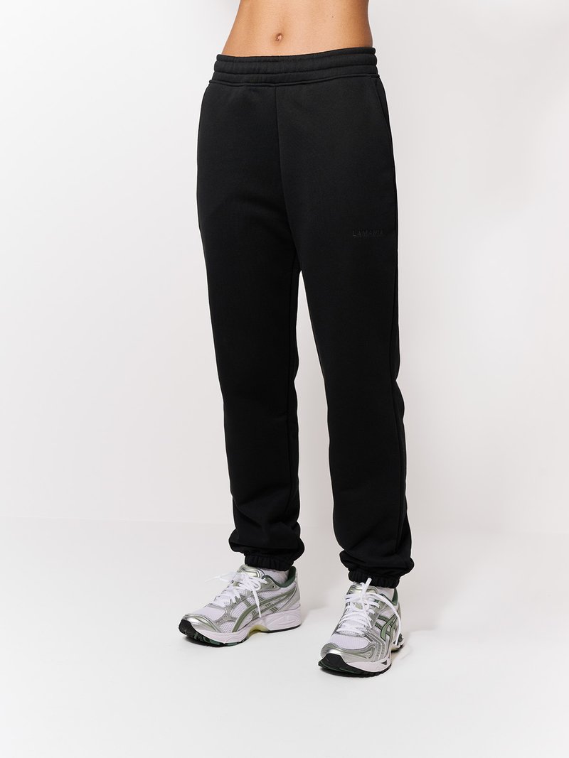Czarne spodnie dresowe z elastycznym pasem i ściągaczami przy kostkach, o gładkiej fakturze i subtelnym logo na nogawce, w połączeniu z szarymi butami sportowymi.