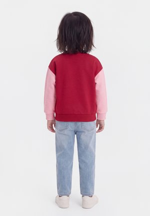 Barn med mellemlangt mørkt hår står med ryggen til og bærer en rød og lyserød langærmet trøje, lyseblå jeans og hvide sko.