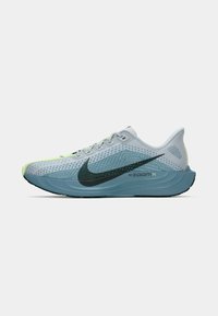 Gaiši zils Nike ZoomX skriešanas apavs ar melnu swoosh, laima zaļiem akcentiem un biezu zilu zoli, attēlots no kreisās puses uz balta fona.