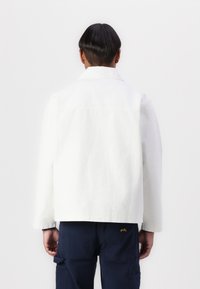 White denim jacket with a classic collar and long sleeves, featuring a relaxed fit and textured fabric. Back view shows simplicity in design.