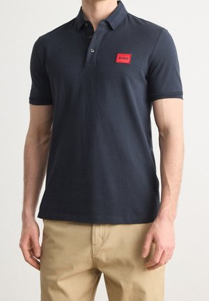 Homme portant un polo bleu marine avec un petit logo rouge "HUGO" sur la poitrine et un pantalon beige, sur fond uni.