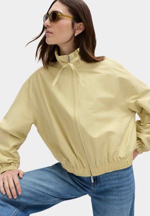 Kvinde iført lys gul lynlåstrøje med snøre ved kraven, blå jeans og solbriller med grønne stel, poserer med det ene knæ løftet.
