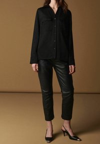 Žena v černé košili na knoflíky, černých kožených kalhotách a černých botách s otevřenou patou a kovovými špičkami, stojící na hnědém pozadí.