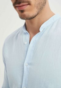 Homme avec une légère barbe portant une chemise à col mandarin bleu clair avec les boutons du haut défaits, sur un fond uni.