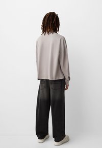 Vaaleanharmaa ylisuuri collegepaita pitkillä hihoilla, yhdistettynä löysiin mustiin gradient-farkkuihin ja valkoisiin lenkkareihin, nähtynä takaa.