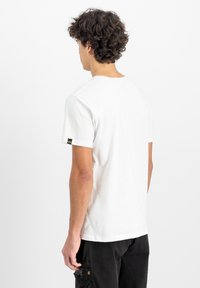 Hvid bomulds t-shirt med korte ærmer, rund halsudskæring og et lille sort logo-patch på ærmet. Glat tekstur og afslappet pasform.