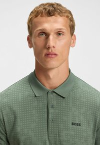 Grön polo t-shirt i strukturerad tyg med rutigt mönster, tre knappar och en svart logotyp på vänster sida.