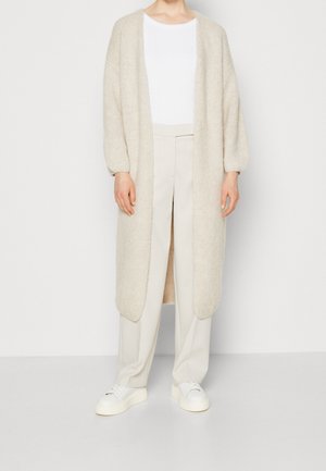 Persona con un cárdigan largo beige de punto, top blanco, pantalones beige de pierna ancha y deportivas blancas, de pie frente a un fondo claro y sencillo.