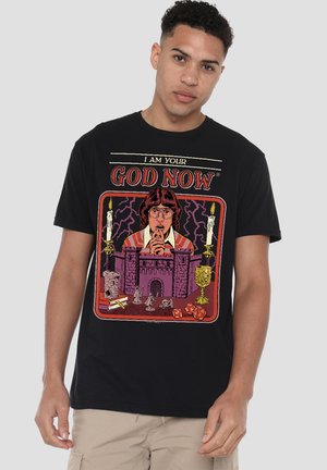 Junger Mann trägt ein schwarzes T-Shirt mit einer Grafik einer betenden Person vor einer Burg, Kerzen, Würfeln und dem Text "Ich bin jetzt dein Gott."