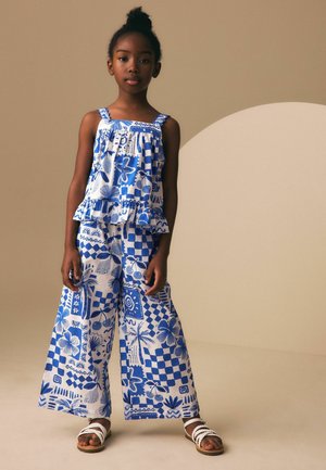 Dziewczyna stoi, ubrana w niebiesko-biały, bezrękawnik w wzory z falbankami oraz pasujące szerokie spodnie, w białych sandałach, na neutralnym tle.