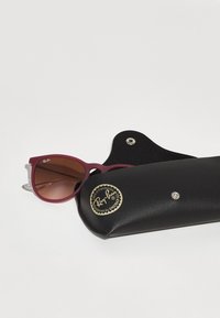 Ochelari de soare cu un cadru din plastic burgund și lentile maro cu un gradient, odihnindu-se într-o husă din piele neagră cu un logo auriu și închidere tip clapetă.