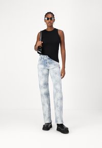 Top sin mangas negro, jeans con estampado de serpiente azul, zapatos de plataforma gruesa negros, modelo sosteniendo una pequeña bolsa negra, fondo neutro.