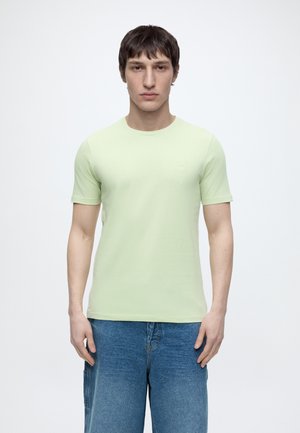 Mężczyzna z krótkimi ciemnymi włosami, ubrany w jasnooliwkowy T-shirt z krótkim rękawem i niebieskie dżinsy, stoi na jednolitym białym tle.