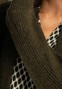 Oliivinvihreä, teksturoitu neulecardigan on drapattu kuviollisen blousen päälle, jossa on vihreitä ja kermaisia muotoja. Lähikuva kankaan rakenteesta ja kerroksellisuudesta.