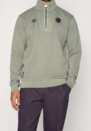Man draagt lichtgroen quarter-zip fleece met twee zwarte logo's op de borst en donkere broek, gedeeltelijk zichtbaar van nek tot halverwege de dij.