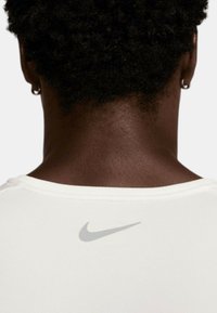 Camisa blanca de Nike con cuello redondo, que presenta un pequeño logo de swoosh en gris en la parte posterior. La tela parece suave y ajustada.