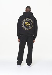 Felpa nera con un grande logo circolare dorato sul retro. Abbinata a pantaloni neri e sneakers bianche, presentata su uno sfondo semplice.