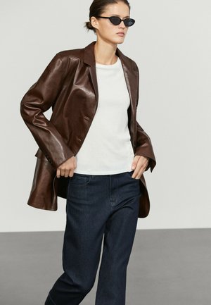 Donna che indossa un blazer in pelle marrone sopra una camicia bianca e jeans scuri, con occhiali da sole neri e capelli legati all'indietro.