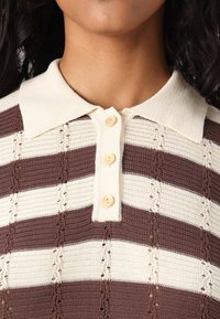 Strikket polo skjorte med brune og kremfargede striper, med krage, tre knapper og dekorative hullmønstre.