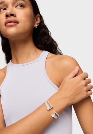 Bracciali in argento con catene d'argento testurizzate, caratterizzati da un design floreale. Indossati al polso sopra una canotta di colore viola chiaro.