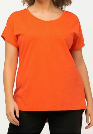 Donna che indossa una maglia arancione brillante a maniche corte con bottoni lungo le cuciture delle spalle e pantaloni neri, in piedi contro uno sfondo semplice.