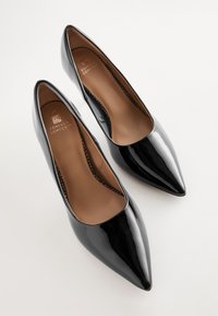Zapatos de tacón alto de charol negro con punta afilada, forro interior marrón y la marca "Forever Comfort" en la plantilla.