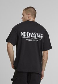Sort bomulds oversized t-shirt med hvid tekst på ryggen: "NO DAYS OFF" og "Another Cotton Sale." Enkel rund halsudskæring, korte ærmer.