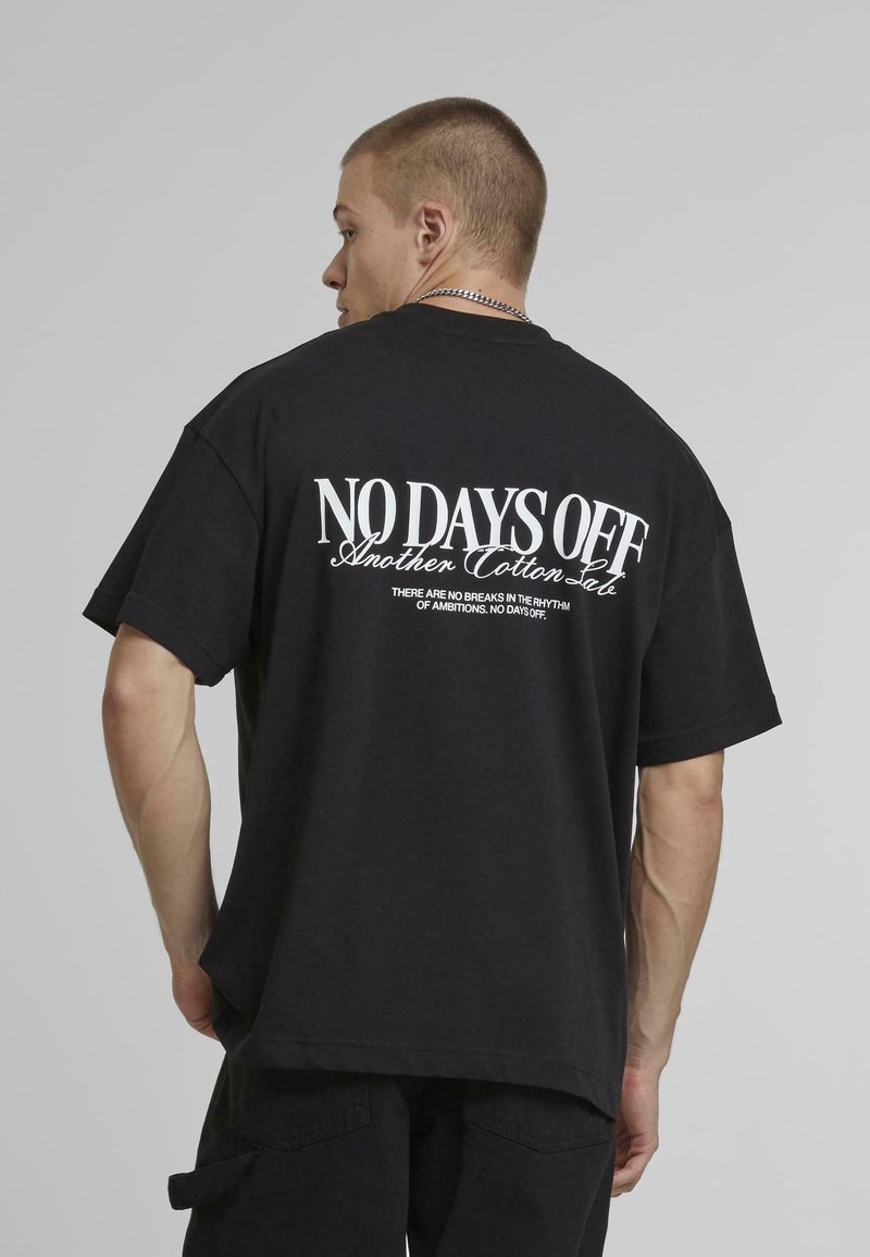 Sort bomulds oversized t-shirt med hvid tekst på ryggen: "NO DAYS OFF" og "Another Cotton Sale." Enkel rund halsudskæring, korte ærmer.