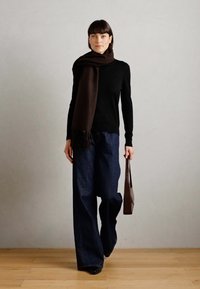 Pull noir, écharpe marron foncé, pantalon en denim bleu foncé ample et chaussures noires brillantes. Porte un sac en cuir marron. Fond uni.
