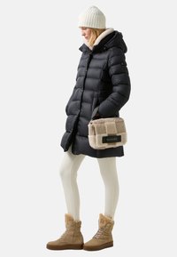 Černá péřovka s kapucí podšitou umělou kožešinou, krémová pletená čepice, béžová kabelka s kožešinovými akcenty a hnědé boty s kožešinovým podšívkou. Bílé legíny.