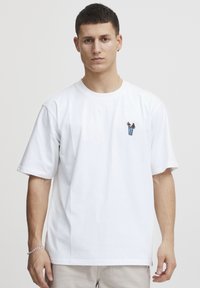 Hvit bomulls t-skjorte med korte ermer, med et lite, fargerikt brodert design på brystet. Klassisk rund hals og avslappet passform.