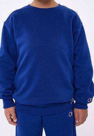 Barn iført en blå Champion sweatshirt og matchende blå bukser, stående med armene afslappet ned langs siderne mod en ensfarvet baggrund.