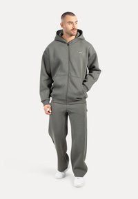 Un homme marchant, portant un sweat à capuche zippé gris et un pantalon de jogging assorti, avec des baskets blanches, regardant sur le côté.