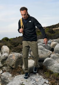 Chaqueta de forro polar negra con cremallera, pantalones cargo caqui y zapatillas oscuras. El sujeto lleva una mochila amarilla y camina por un terreno rocoso con arbustos.