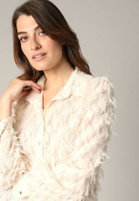 Vrouw met lang bruin haar in een textuur blouse in crème, die haar kraag aanraakt, neutrale achtergrond, zachte belichting.