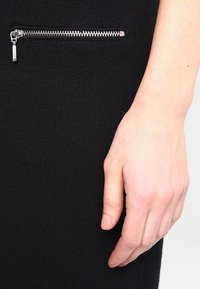 Prenda de punto negra con un bolsillo con cremallera y textura acanalada. La cremallera es plateada, lo que añade un detalle funcional al diseño.