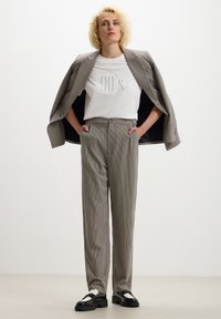 Blazer grigio a scacchi con pantaloni a vita alta coordinati, abbinato a una camicia bianca con la scritta "90's" in lettere testurizzate e scarpe nere.
