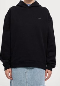 Felpa nera con un taglio largo, realizzata in un tessuto morbido. Presenta una tasca frontale e un piccolo logo sul petto. Abbinata a jeans in denim chiaro.