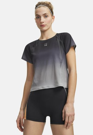 Young woman wearing black and gray gradient short-sleeve athletic shirt and black fitted shorts standing against a white background.