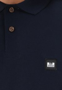 Navy blue polo shirt made of knit fabric, featuring a collar, brown buttons, and a black rectangular logo patch with "WEEKEND OFFENDER."