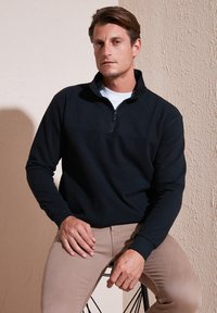 Felpa nera con zip, maniche lunghe e colletto alto, abbinata a pantaloni beige. Tessuto liscio, design casual. Semplice e funzionale.