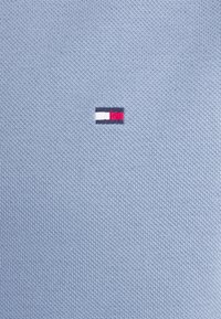 Petit logo rectangulaire avec des sections horizontales blanches, rouges et bleu foncé brodées sur un tissu texturé bleu clair.