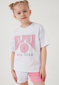 Jeune fille aux cheveux tressés portant un T-shirt blanc oversize avec l'inscription "ALPHA GIRL TEAM" en rose et gris, et un short rose et blanc.