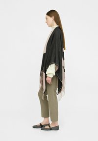 Poncho grigio e rosa con frange, indossato sopra un maglione bianco, abbinato a pantaloni beige e ballerine nere con motivo, mostrato in vista di profilo.