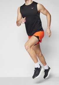 Černé bezrukávové sportovní tričko, oranžové šortky s černými akcenty a černé běžecké boty s bílou podrážkou, model v běžecké pozici.